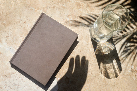Book and Glass of Water on Table. A beige book and a clear glass of water casting shadows on a light brown tiled surface.の写真素材