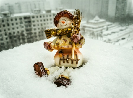 A toy snowman sitting in snow, snowfall cityscapeの素材