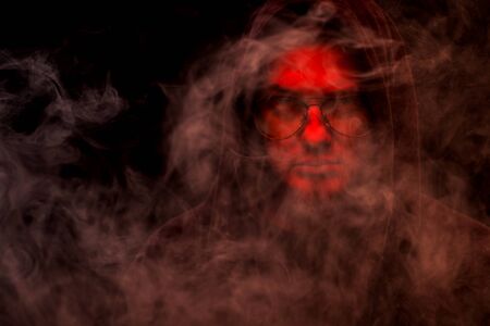 A warlock with a red face in a hood and glasses on a black background in smoke.の写真素材