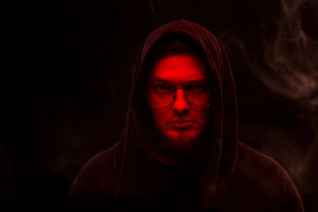 A warlock with a red face in a hood and glasses on a black background in smoke.の写真素材