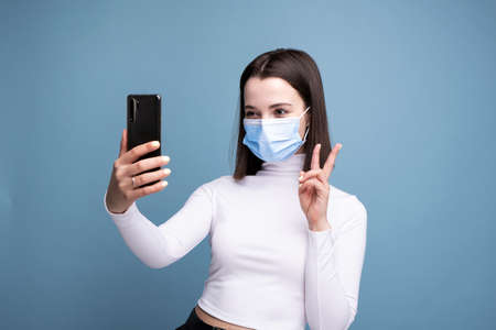 A young woman in a medical mask holds a mobile phone in her hands and makes a selfie on a blue background in the studio.の写真素材
