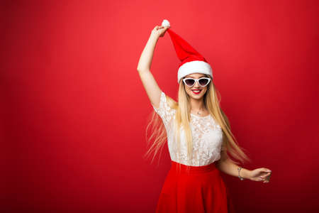 Happy blonde in santa hat on red isolated background. White rimmed sunglasses.の写真素材