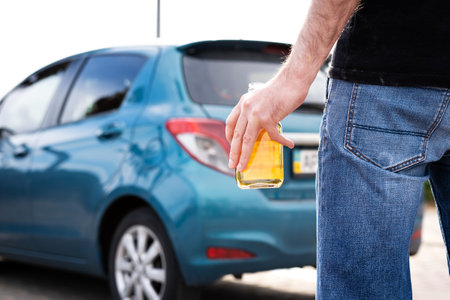 A man in jeans with a bottle of whiskey in his hand tries to get behind the wheel of a car.の写真素材
