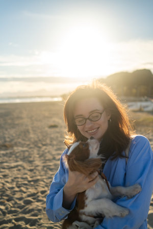 A young woman hugs her Cavalier King Charles Spaniel on the beach at sunset. Warm light, waves, and smiles capture a peaceful, joyful moment of connection and relaxation.の写真素材