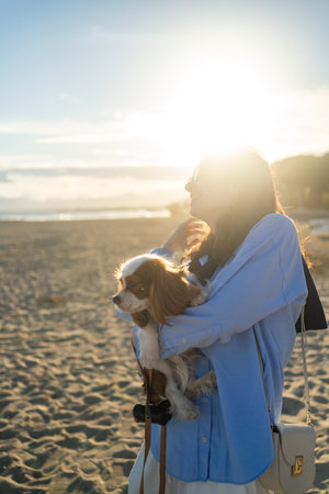 A young woman hugs her Cavalier King Charles Spaniel on the beach at sunset. Warm light, waves, and smiles capture a peaceful, joyful moment of connection and relaxation.の写真素材