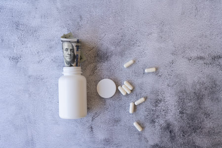 White pill bottle filled with dollar bills and scattered capsules on a gray background, concept of...