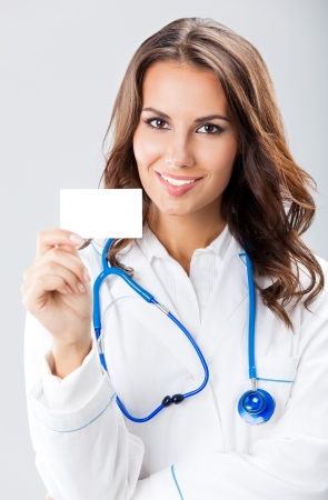 Portrait of happy smiling young female doctor showing blank business card or invitation, over grey backgroundの写真素材