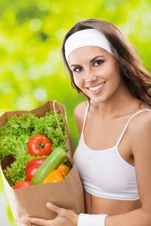 Portrait of happy smiling young beautiful woman in fitness wear holding grocery shopping bag with healthy vegetarian food, outdoorsの写真素材