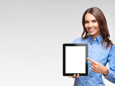 Happy smiling beautiful young businesswoman showing blank no-name tablet pc monitor, against grey background, with copyspace areaの写真素材