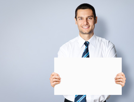 Portrait of happy smiling young businessman showing blank signboard, with copyspace area for text or slogan, against grey backgroundの写真素材
