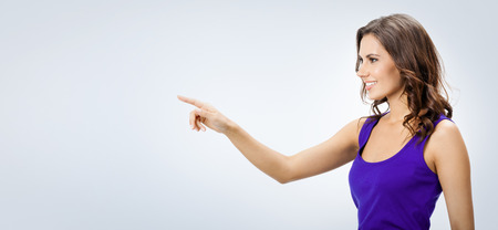 Cheerful beautiful young woman in violet casual smart clothing, showing copyspace, visual imaginary or something, or pressing virual button, over grey background, with blank area for slogan or product or textの写真素材