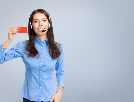 Portrait of smiling cheerful customer support phone operator in headset, showing red blank business or plastic card, against grey backgroundの写真素材