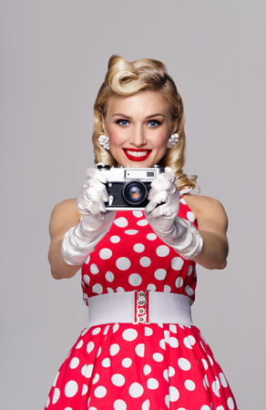 Portrait of beautiful young happy smiling woman, with no-name old film camera, taking picture, dressed in pin-up style. Caucasian blond model posing in retro fashion and vintage concept studio shoot, on grey background.の写真素材