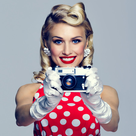Portrait of beautiful young happy smiling woman, with no-name old film camera, taking picture, dressed in pin-up style. Caucasian blond model posing in retro fashion and vintage concept studio shoot.の写真素材