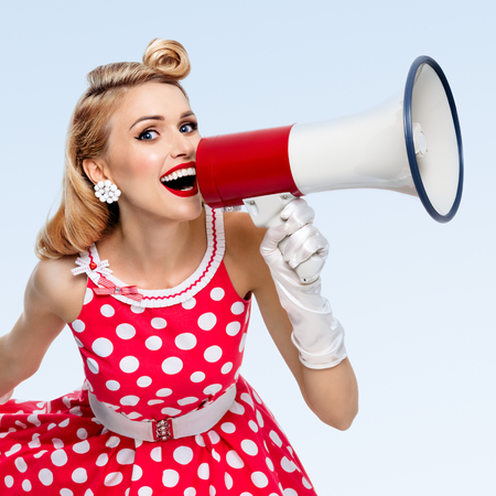 Portrait of woman holding megaphone, dressed in pin-up style red dress in polka dot and white gloves, on blue background. Caucasian blond model posing in retro fashion vintage studio shoot.の写真素材