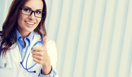 Happy smiling young female doctor in glasses, showing thumbs up hand sign gesture, at office, with blank copyspace area for slogan or advertise text. Healthcare, medical, lab consulting and exam concept.の写真素材