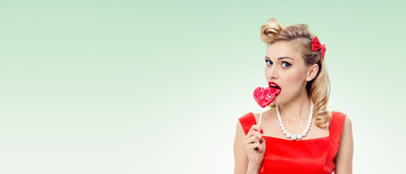 Woman eating heart shape lollipop dressed in pinup style dress in polka dot, on green background. Caucasian blond model posing in retro fashion and vintage concept studio shoot. Copyspace area for advertising slogan or text message.の写真素材