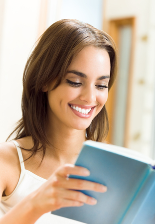 Portrait of young happy smiling beautiful woman reading a book or textbook at homeの写真素材