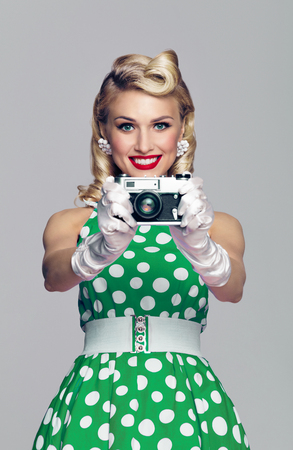 Portrait of beautiful young happy smiling woman, with no-name old film camera, taking picture, dressed in pin-up style. Caucasian blond model posing in retro fashion and vintage concept studio shoot, on grey background.の写真素材