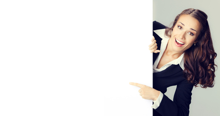 Portrait of happy smiling young woman in black confident clothing showing blank signboard with copyspace empty area for some text or slogan, over grey background. Success in business and advertising concept photo.の写真素材