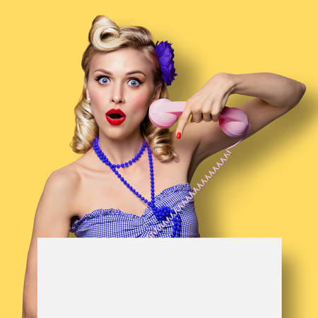 Excited surprised woman with phone and blank signboard. Pin up girl. Blond model at retro fashion and vintage concept. Yellow color background. Copy space for some advertise slogan, imaginary or text.の写真素材