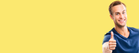 Portrait of happy smiling man in blue casual clothing, showing thumb up gesture, over yellow color...