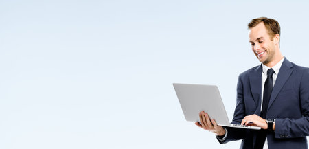 Smiling young successful businessman in black confident suit, using laptop pc or notebook, over grey background. Success in business concept.の写真素材