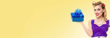 Retro, vintage  and holiday sales concept picture - happy pinup style lovely girl, holding gift box, with copy space for some slogan or advertising text, over yellow background.の写真素材