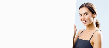 Happy smiling young fit beautiful woman in black fitness wear showing blank signboard or banner with copy space for some slogan or text, over grey backgroundの写真素材