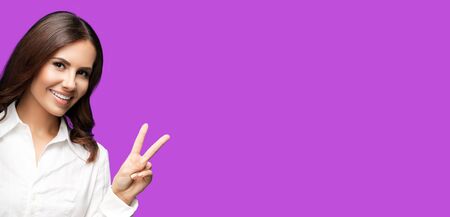 Portrait of happy smiling young cheerful confident businesswoman, showing two fingers or victory hand gesture, over violet purple background. Success in business concept picture.の写真素材
