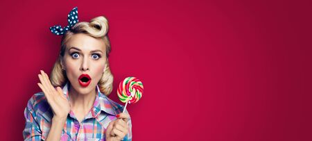 Excited woman with lollipop. Girl pin up with open mouth. Blond model at retro fashion and vintage concept. Dark red color background. Copy space for some text . Wide horizontal banner composition.の写真素材