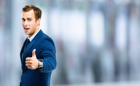 Excited businessman showing thumbs up like hand sign gesture, in blue confident suit, blurred office...