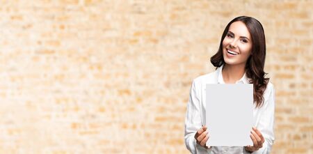 Portrait of happy smiling brunette businesswoman showing blank signboard with copy space area for some slogan or text, over loft style wall background. Success in business concept.の写真素材