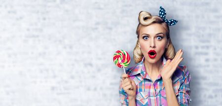 Excited surprised woman with lollipop. Girl pin up with open mouth. Blond model at retro fashion and vintage concept. White brick wall background. Copy space for some advertise slogan, sign or text.の写真素材