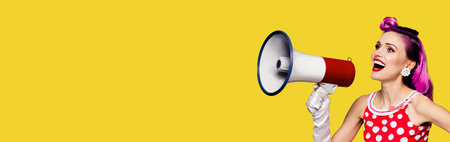 Purple head woman using megaphone, shouting something. Pin up girl in red polka dot dress. Caucasian model posing in retro vintage concept. Yellow color background. Wide composition with copy space.の写真素材