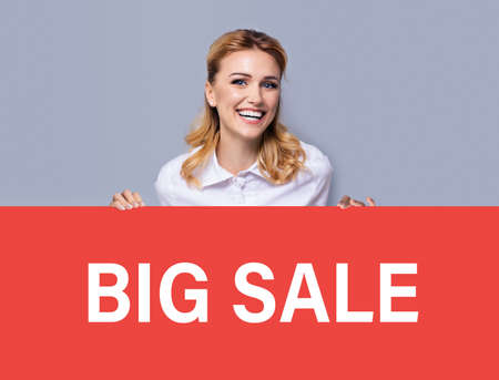 Very happy excited, laughing business woman in white confident clothing showing red banner signboard with big sale sign. Advertising studio concept. Blond girl standing behind broadsheet, over grey.の写真素材