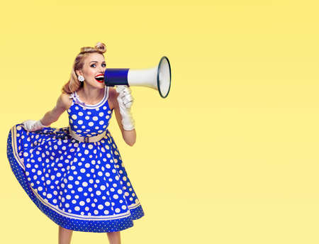 Happy excited woman holding megaphone, dressed in pin up style blue dress in polka dot and white gloves, on yellow background. Beautiful blond girl in retro fashion vintage studio picture.の写真素材