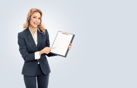 Call center. Businesswoman or female support phone operator in headset, showing blank clipboard with copy space for some text, isolated over grey background. Customer service help consulting concept.の写真素材