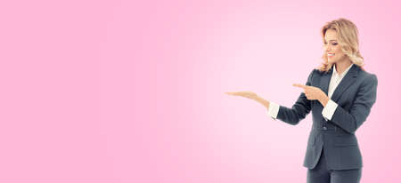 Smiling businesswoman in grey confident suit, showing  or holding something, some product or blank copy space for text, isolated over pink color background.の写真素材