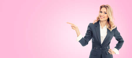 Happy smiling businesswoman in grey confident suit, pointing at something, some product or blank copy space area for text, isolated on pink color background.  Business studio concept.の写真素材