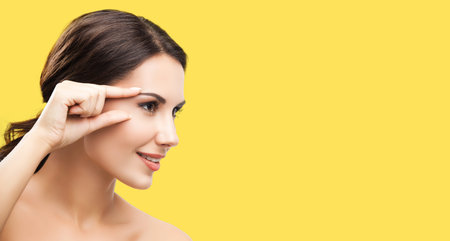 Skin care beauty concept. Portrait image of smiling woman pointing brow or eye, touching skin, applying face cream, isolated over yellow color background. Brunette model at studio.の写真素材