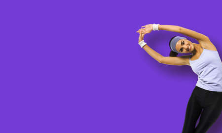 Happy smiling african american woman in sportswear doing stretching fitness exercise or yoga moves, isolated over violet purple background. Sporty girl at studio. Copy space empty area for some textの写真素材
