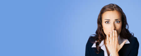 Surprised excited young businesswoman in confident black suit and green blouse, covering with hand her mouth, copy space place for some text or slogan, over blue color background. Business ad concept.の写真素材