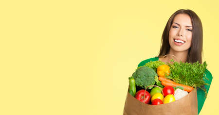 Woman holding shopping bag with healthy vegetarian raw food, copy space area for text, isolated against yellow color background. Portrait image of asian caucasian girl at studio.の写真素材