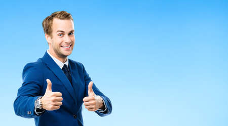 Portrait of confident businessman showing thumbs up hand sign gesture, on blue background. Business success concept. Young happy smiling man at studio.の写真素材
