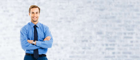 Portrait of happy smiling businessman, standing in crossed arm pose, over white brick loft wall background. Confident business man. Copy space area.の写真素材