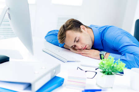 Image of very tired businessman in blue shirt, sleeping at workplace, with computer, indoors. Business, job and overworking concept.の写真素材