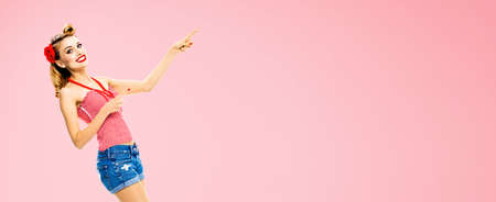 Portrait image of amazed happy woman pointing at something. Excited girl in pin up style, showing copy space for text or imaginary. Retro fashion and vintage. Rose pink colour back.の写真素材