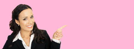 Happy smiling attractive woman in black confident suit, pointing showing copy space area. Business concept. Rose pink colour background. Brunette businesswoman. Wide image.の写真素材