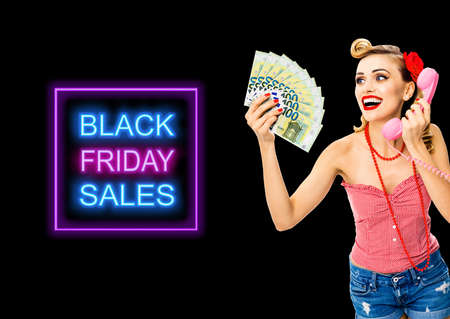 Happy excited astonished woman holding money euro cash banknotes, talking on phone, dressed in pin up style. Blond girl in retro fashion and vintage ad concept. Black Friday sales neon light sign textの写真素材